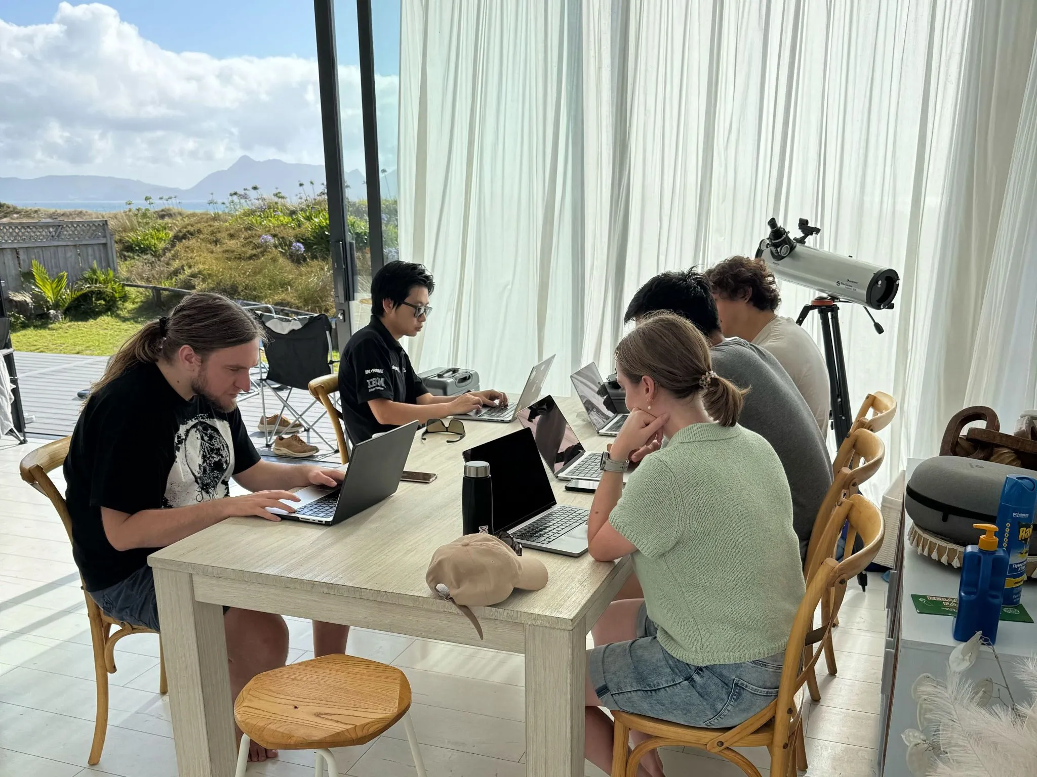 Five team members heads-down on laptops at a shared table, ocean visible through the window